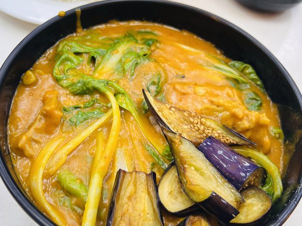 Large bowl of kare kare with eggplant, string beans and pechay in thick peanut sauce, served with steamed rice and a side of bagoong.