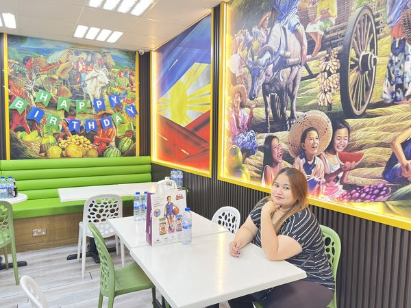 Wide view of the dining room showing Filipino harvest murals, a Philippine flag lightbox, green booth seating and white tables, with a woman seated at one table.