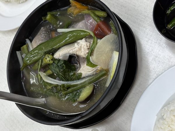 Overhead flat lay of sinigang na isda in a black bowl with fish, okra, string beans, radish, green chilli and kangkong in tamarind broth, with side bowls of broth, plates of rice and lemon wedges.