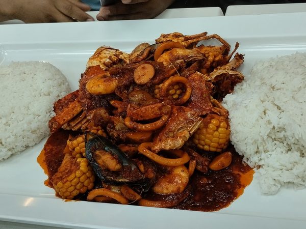 Long white platter of a spicy seafood boil with squid rings, shrimp, mussels, crab, corn and sausage in red sauce, flanked by two mounds of steamed rice.