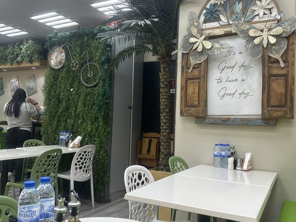 Interior of the restaurant with white tables, green chairs, a green foliage wall with a vintage clock and bicycle decor, and a framed Good day wall sign.