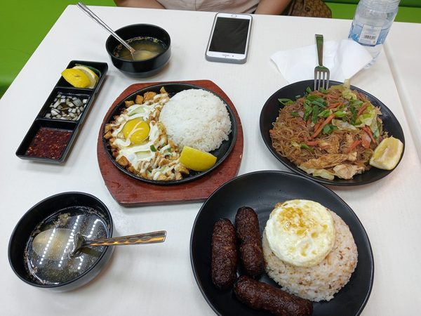 Overhead spread of sizzling sisig with a raw egg and rice on a hot plate, pancit bihon, longganisa with garlic rice and fried egg, two soup bowls and a condiment tray.