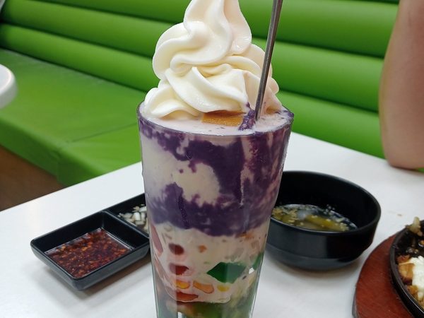 Tall glass of halo halo layered with ube, leche flan and coloured jellies under a swirl of soft serve, on a table beside a bowl of soup and a sizzling plate.