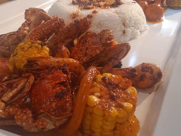 Seafood boil platter of crab, prawns and corn on the cob in a red garlic-butter sauce, served with a mound of garlic rice on a white plate