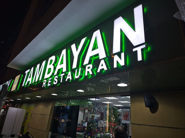 Illuminated Pinoy Tambayan Restaurant shopfront sign at night, with a Christmas tree and Merry Christmas decorations visible through the glass