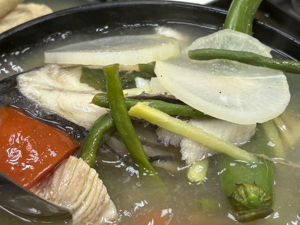 Bowl of fish sinigang in sour tamarind broth with radish slices, tomato, long beans and green chilli, with steamed rice behind.