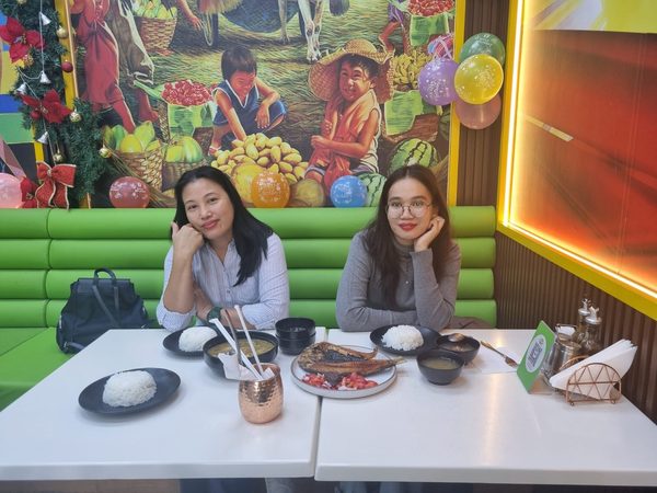 Two women posing in a green booth in front of the Filipino market mural, with grilled bangus on tomato and onion salsa, bowls of soup and plates of steamed rice on the table