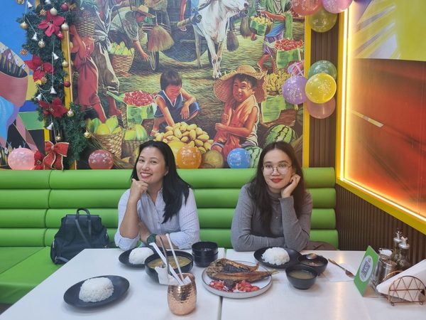 Wider view of two women posing in a green booth under the market mural and birthday balloons, with grilled bangus, soup and steamed rice laid out in front of them