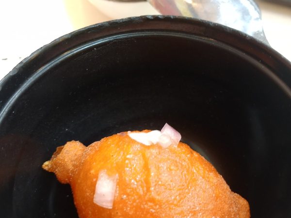 Deep-fried orange-battered egg, tokneneng, in a black bowl scattered with chopped red onion, with a dish of dipping sauce behind